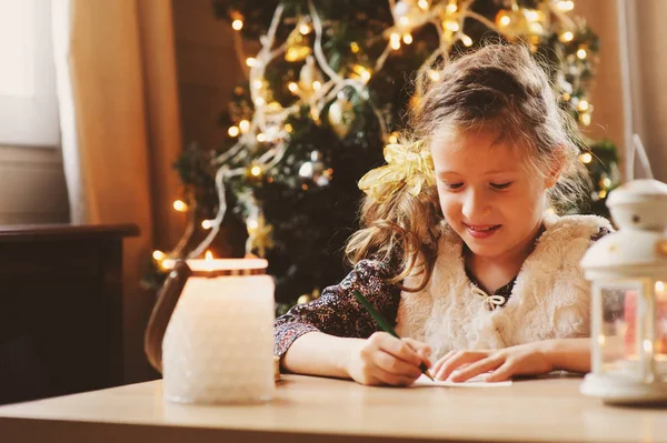Niña escribiendo carta a Santa en casa. Niña de 8 años haciendo lista de regalos para Navidad o Año Nuevo en casa con árbol decorado en el fondo — Foto de Stock