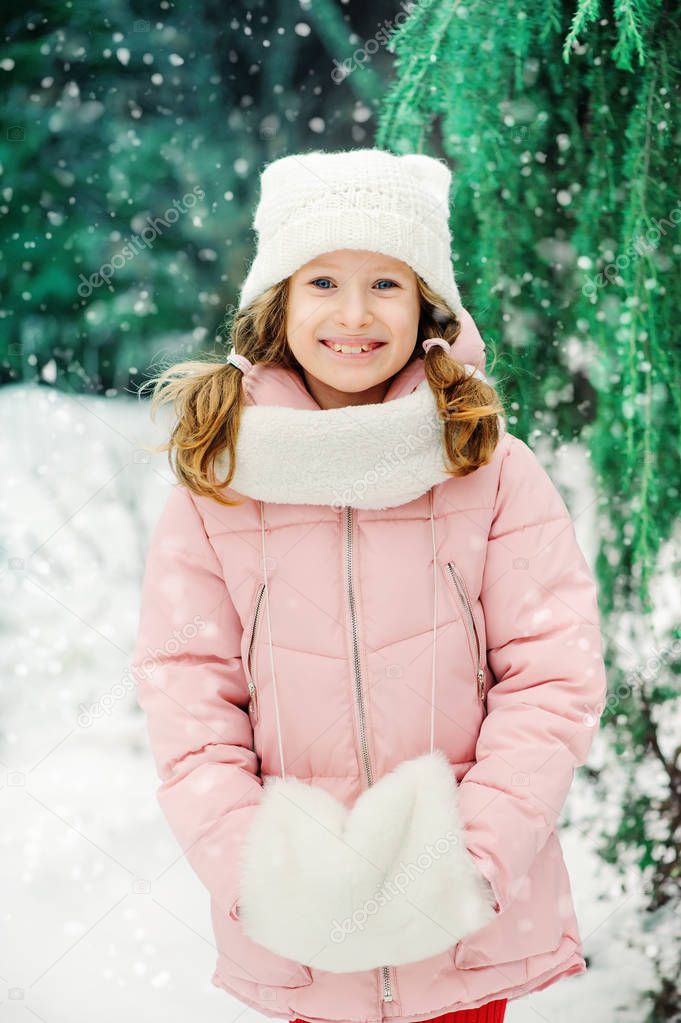 winter portrait of 8 years old kid girl walking outdoor in snowy day, wearing white knitted hat and pink coat