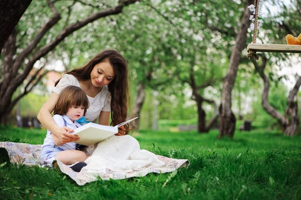 loving mother reading book to toddler son outdoor on picnic in spring or summer park. Happy family and mothers day concept