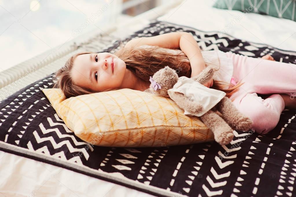 happy little child girl lying on her bed in the morning, waking up in comfortable room with modern bedlinen and pillows, cozy homely scene 