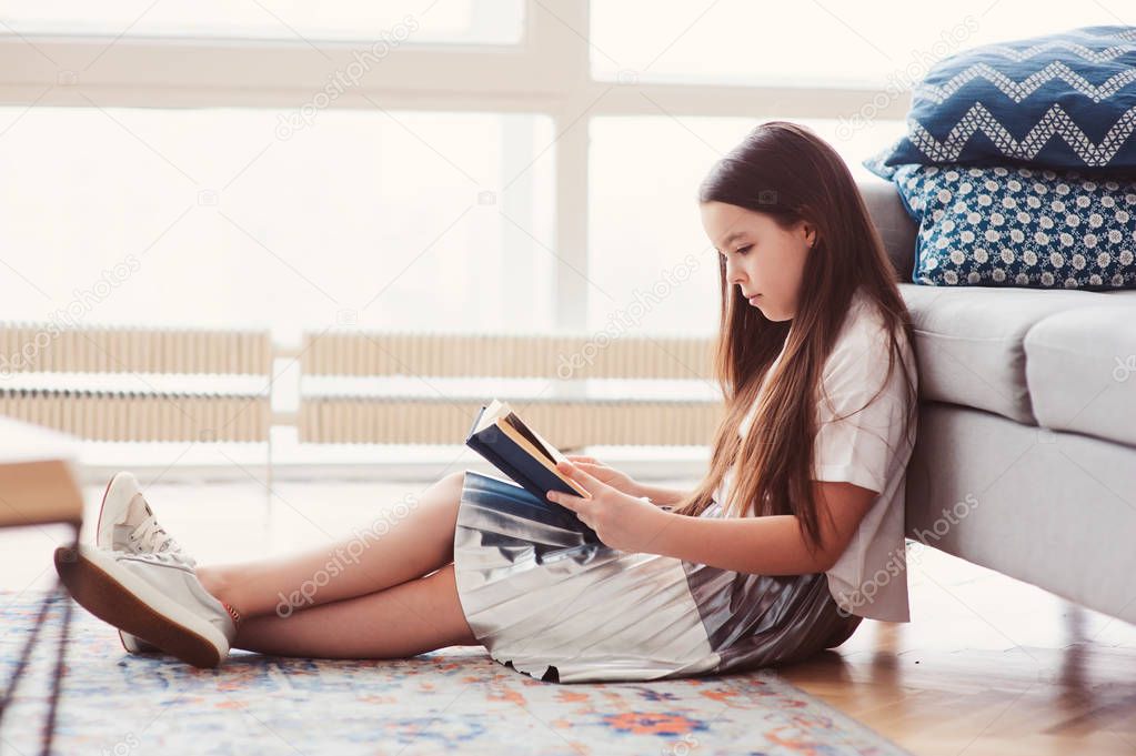 smart kid girl reading interesting book  at home, sitting with couch on background. Learning and education concept