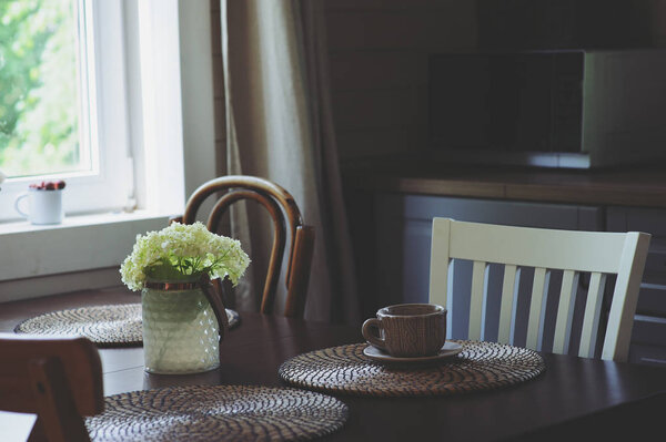 cozy summer morning at rustic country house kitchen. Wooden table with bouquet of fresh flowers, open shelving. Casual breakfast in grey and brown interior.