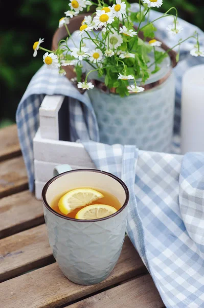 Summer outdoor breakfast with tea, lemon and bouquet of chamomile flowers on wooden table. Summertime relax and country living concept