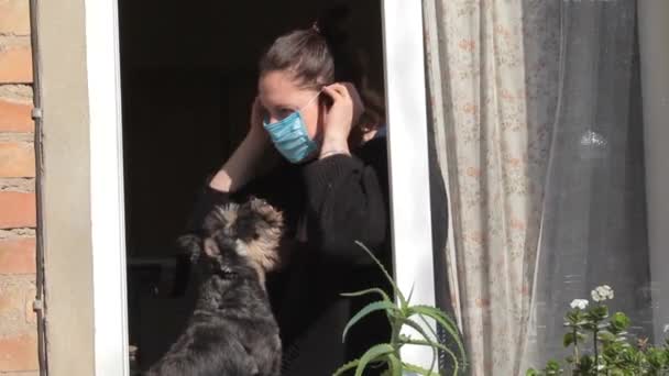 Självisolering, coronavirusepidemi. Kvinna i skyddsmask med hund tittar genom fönstret. Karantän för Coronavirus och sällskapsdjur — Stockvideo