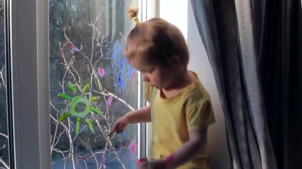 Toddler lány fest az ablakon, és mutatja a kezét a festékben. Kölyök otthon. Karantén — Stock videók