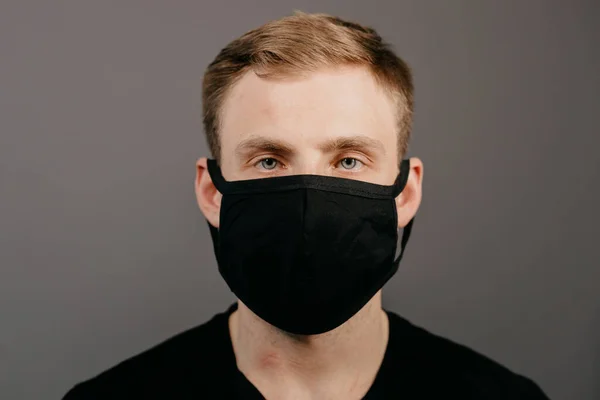 Hezký muž v černém tričku nosí černou lékařskou masku, šedé pozadí. Koronavirus covid-19 koncepce období karantény — Stock fotografie