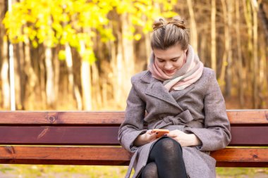 Paltolu genç ve çekici bir kadın bankta oturuyor ve sonbahar parkında telefonla bir mesaj okuyor.