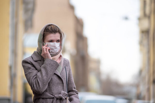 Young attractive European Caucasian woman in a medical protective mask against influenza viruses and coronavirus COVID-19 talking on the phone on a city street