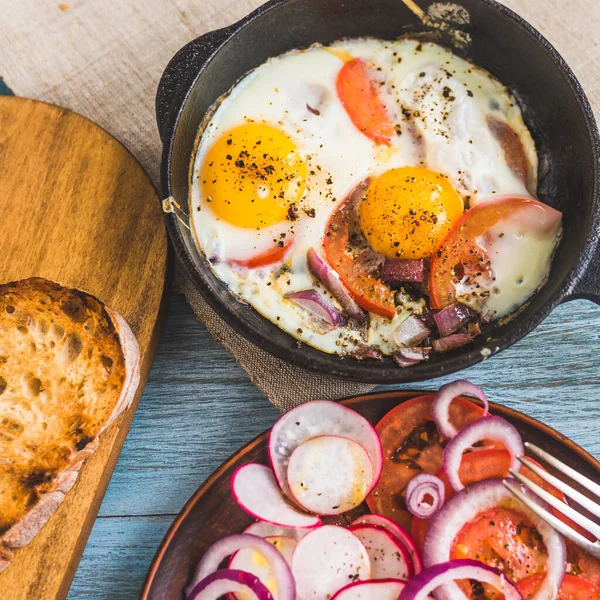 Rustic breakfast in a family hotel - scrambled eggs with vegetables in a cast-iron pan and fresh salad with radish, tomatoes and red onion rings in a plate, top view, close-up