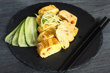 Geleneksel Japon omleti Tamagoyaki ve çubuklu siyah tabakta dilimlenmiş salatalık.