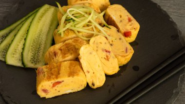 Geleneksel Japon omleti Tamagoyaki ve çubuklu siyah tabakta dilimlenmiş salatalık.