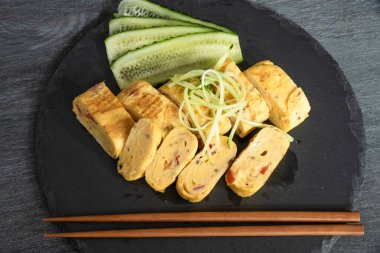 Geleneksel Japon omleti Tamagoyaki ve siyah tahta üzerinde salatalık dilimlenmiş yemek çubukları, üst manzara