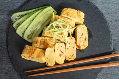 Geleneksel Japon omleti Tamagoyaki ve siyah tahta üzerinde salatalık dilimlenmiş yemek çubukları, üst manzara