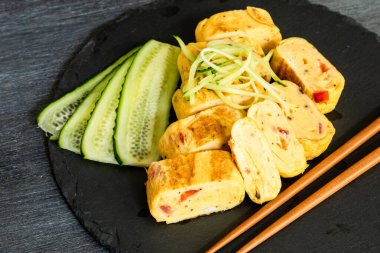 Geleneksel Japon omleti Tamagoyaki ve siyah tahta üzerinde salatalık dilimlenmiş yemek çubukları, üst manzara