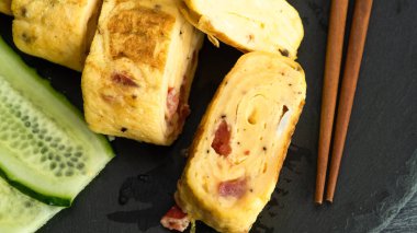 Geleneksel Japon omleti Tamagoyaki ve siyah tahta üzerinde salatalık dilimlenmiş yemek çubukları, yakın çekim