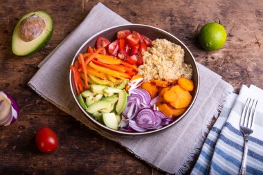 Bir kase kinoalı salata, havuç, avokado, soğan, biber, ahşap bir masada keten peçetede limon dilimleri olan domates.