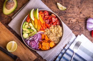 Bir kase kinoalı salata, havuç, avokado, soğan, biber, ahşap bir masada keten peçetede limon dilimleri olan domates.