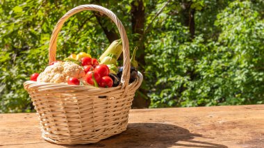 Güneşli bir günde, bahçedeki ağaçların arka planında, ahşap bir masada olgun sebzelerle dolu sepet, yakın plan, kopyalama alanı.