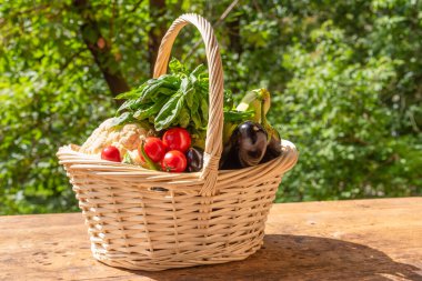 Olgun sebzelerle dolu sepet, bahçedeki ağaçların arka planında, ahşap bir masanın üzerinde.