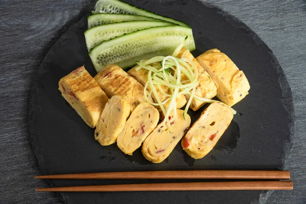 Geleneksel Japon omleti Tamagoyaki ve siyah tahta üzerinde salatalık dilimlenmiş yemek çubukları, üst manzara