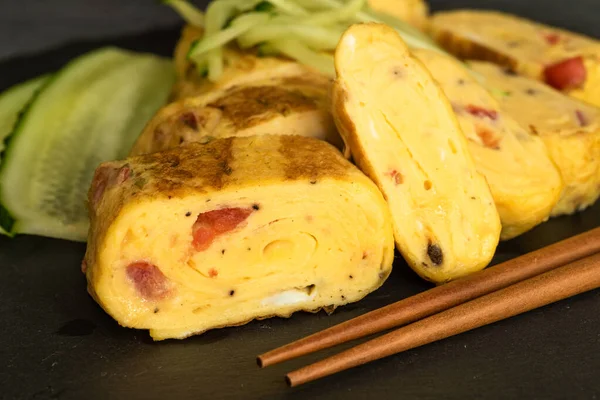 Geleneksel Japon omleti Tamagoyaki ve siyah tahta üzerinde salatalık dilimlenmiş yemek çubukları, yakın çekim
