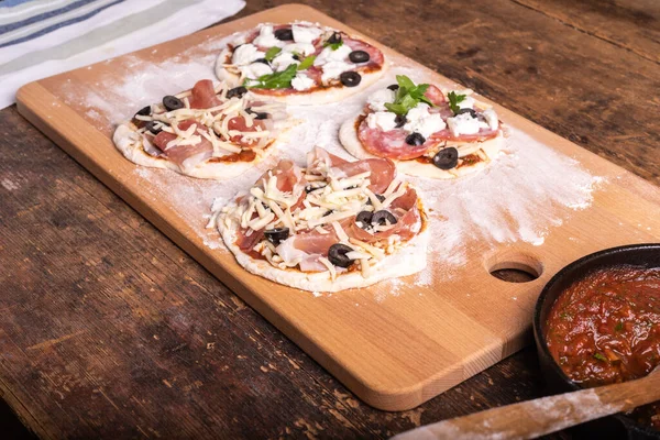 Cooking traditional Italian mini pizza, pizzetta - mini pizza with tomato sauce, salami, jamon and mozzarella cheese on the kitchen blackboard floured