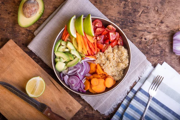 Bir kase kinoalı salata, havuç, avokado, soğan, biber, ahşap bir masada keten peçetede limon dilimleri olan domates.