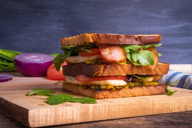 Lezzetli bir köy sandviçi. Tavuklu kızarmış ekmek, kızarmış pastırma, domates, soğan, hardallı turşu ve ahşap tahta üzerinde roka salatası.