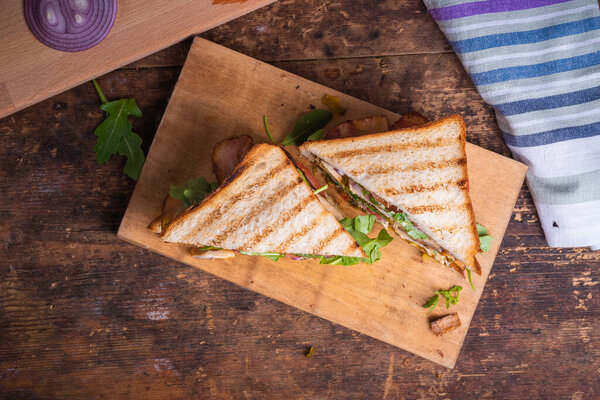 Вкусный сэндвич, разрезанный на треугольники - поджаренный хлеб с куриной грудкой, жареный бекон, помидоры, лук, огурцы с горчицей и салат из рукколы крупным планом на деревянной доске на деревенском столе, вид сверху
