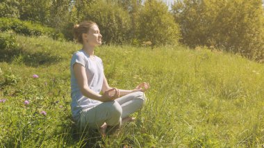 Nilüfer pozisyonunda oturan genç çekici bir kadın ağaçların yakınındaki bir çayırda - doğada dinlenme ve rahatlama kavramı