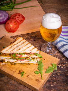 Tavuk göğsü, pastırma, salata ve sebzeli büyük lezzetli ev yapımı üçgen sandviç ve kırsal ahşap arka planda bir bardak bira.
