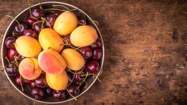 Tahta masadaki bir kasede olgun kayısılar ve kirazlar. Üst manzara, panoramik arka plan ve fotokopi alanı.