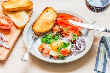 Taze köy kahvaltısı. Domuz pastırmalı, soğanlı ve domatesli sahanda yumurta. Bir bardak kırmızı şarap ve açık ahşap zemin üzerinde kızarmış ekmek.