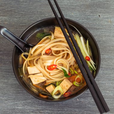 Japon vejetaryen çorbası udon eriştesi, tofu ve sebzeler siyah bir kasede kaşık ve çubuklarla, üst manzara, yakın çekim.