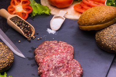 Taze et burgerleri, susamlı çörekler, domates, salata ve baharatlar, arka planda hamburger pişirmek için.