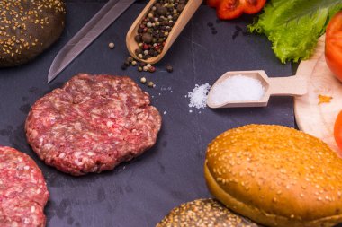 Taze et burgerleri, susamlı çörekler, domates, salata ve baharatlar, arka planda hamburger pişirmek için.