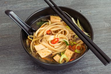 Japon vejetaryen çorbası udon eriştesi, tofu ve sebzeler siyah bir kasede kaşık ve çubuklarla, üst manzara, yakın çekim.