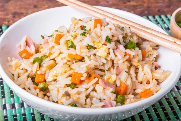 Fried rice with egg, ham and vegetables - a traditional Asian dish of fried rice, eggs, ham, carrots and spices in a bowl on a bamboo napkin, chopsticks, close-up