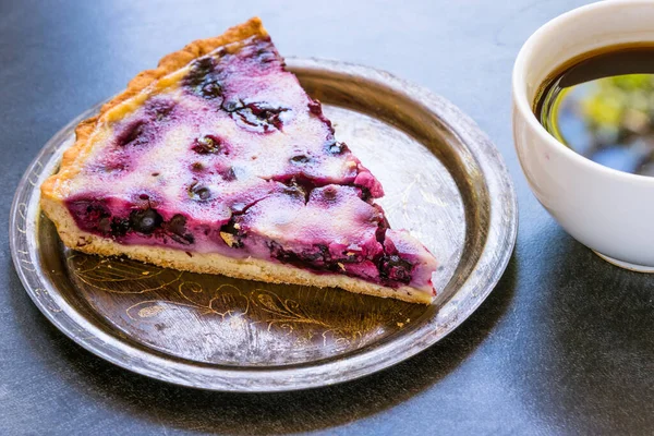 Piece of homemade tart with black currant berries and a cup of coffee on a dark background