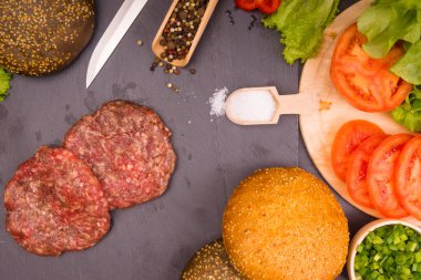 Taze et burgerleri, susamlı çörekler, domates, salata ve baharatlar, arka planda hamburger pişirmek için.
