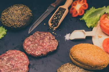 Taze et burgerleri, susamlı çörekler, domates, salata ve baharatlar, arka planda hamburger pişirmek için.