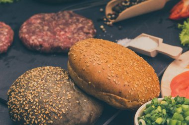 Taze et burgerleri, susamlı çörekler, domates, salata ve baharatlar, arka planda hamburger pişirmek için.