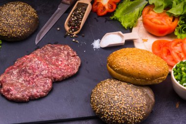 Taze et burgerleri, susamlı çörekler, domates, salata ve baharatlar, arka planda hamburger pişirmek için.
