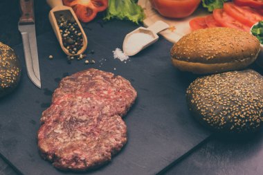 Taze et burgerleri, susamlı çörekler, domates, salata ve baharatlar, arka planda hamburger pişirmek için.