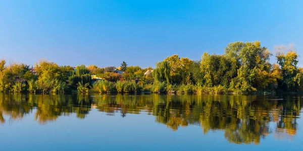 Panoramic autumn landscape - a village on the shore of the lake, houses with trees are reflected in the water