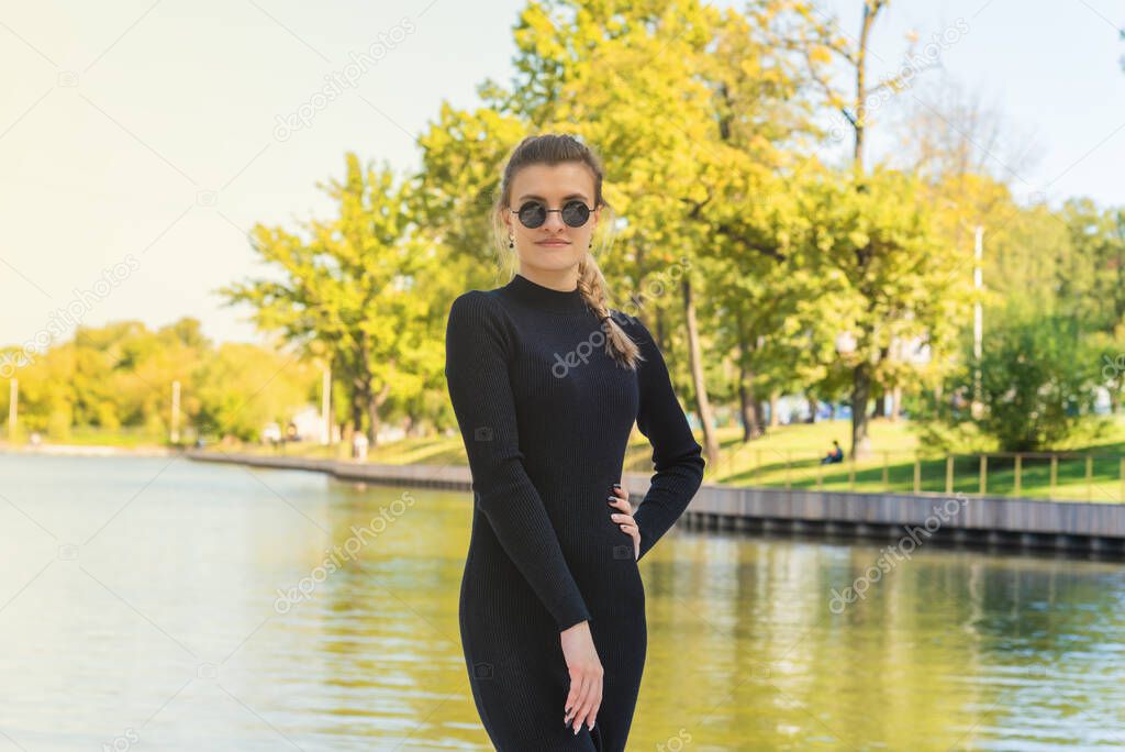 Young attractive slim woman in a tight-fitting black dress and sunglasses. Autumn park with a pond in the background. Beautiful fashionable woman on a park background.