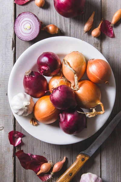 Varios tipos de diferentes bulbos de cebolla en un plato blanco, de pie sobre una vieja mesa de madera . — Foto de Stock