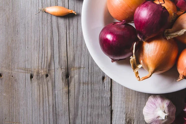 Verschiedene Zwiebelsorten auf einem weißen Teller, der auf einem alten Holztisch steht. — Stockfoto