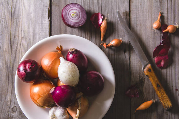 Несколько видов луковых луковиц на белой тарелке, стоящих на старом деревянном столе
.