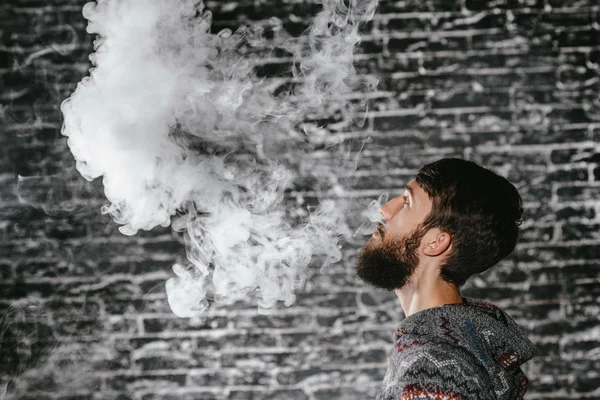 Młody człowiek z brodą wymiotujący elektronicznym papierosem. Vaper hipster dymu parownik. — Zdjęcie stockowe